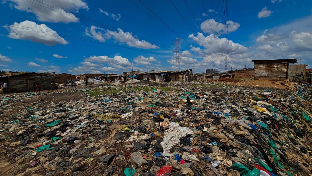 Scattered garbage across an informal settlement with makeshift structures and electrical transmission towers under a bright sky.