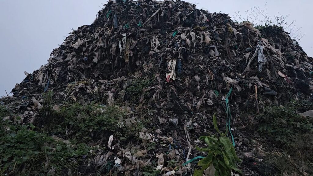 owering mound of decomposing garbage with scattered plastic and fabric waste, surrounded by sparse vegetation under an overcast sky.